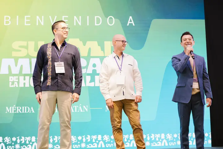 Francisco welcomes the audience with a microphone beside two fellow organizers on the Summit Mexicano de la Hospitalidad stage.